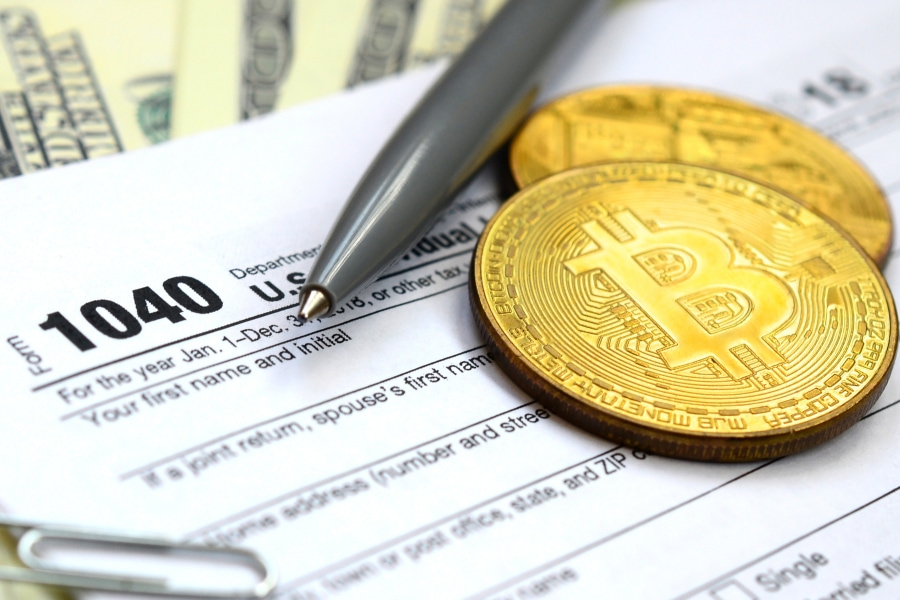 A pen laying on a virtual currency transaction tax form next two physical Bitcoin coins